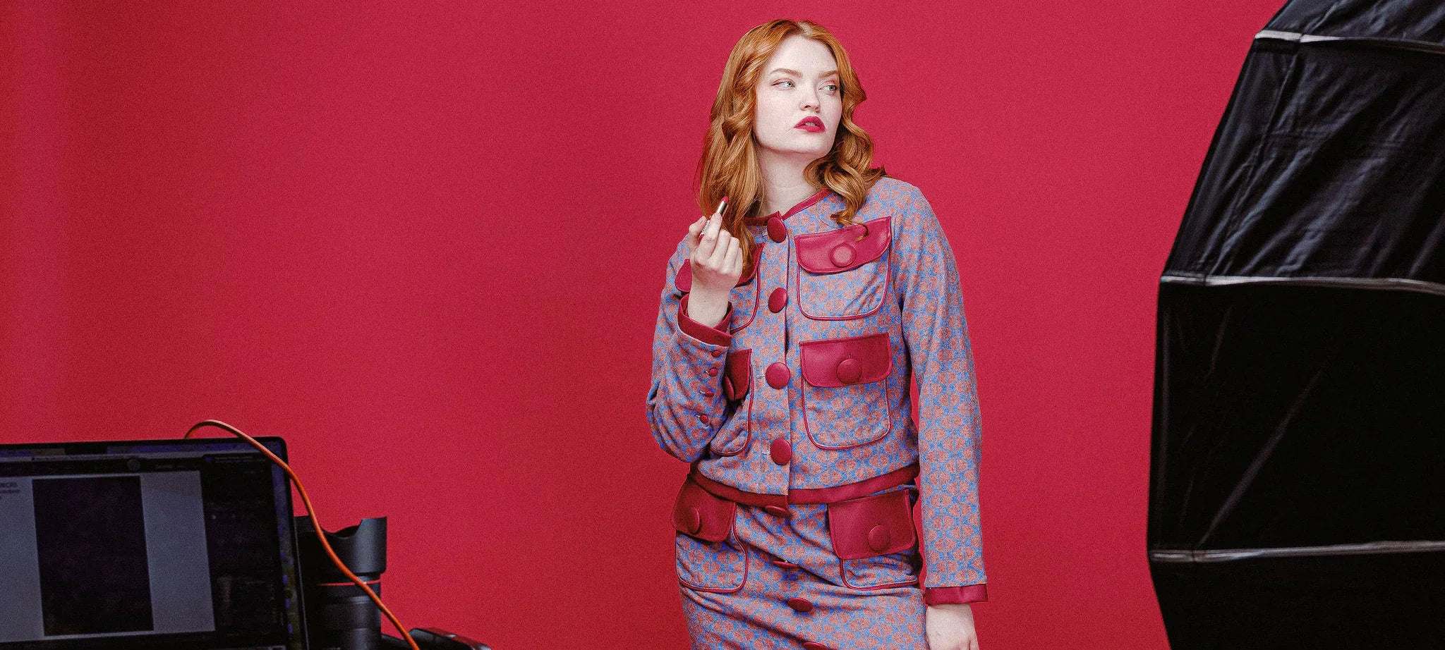 Model wearing the Carly Cardigan Jacket and Carly Mini Skirt from House of Caswell, styled against a bold red backdrop, holding a lipstick in a studio setting.
