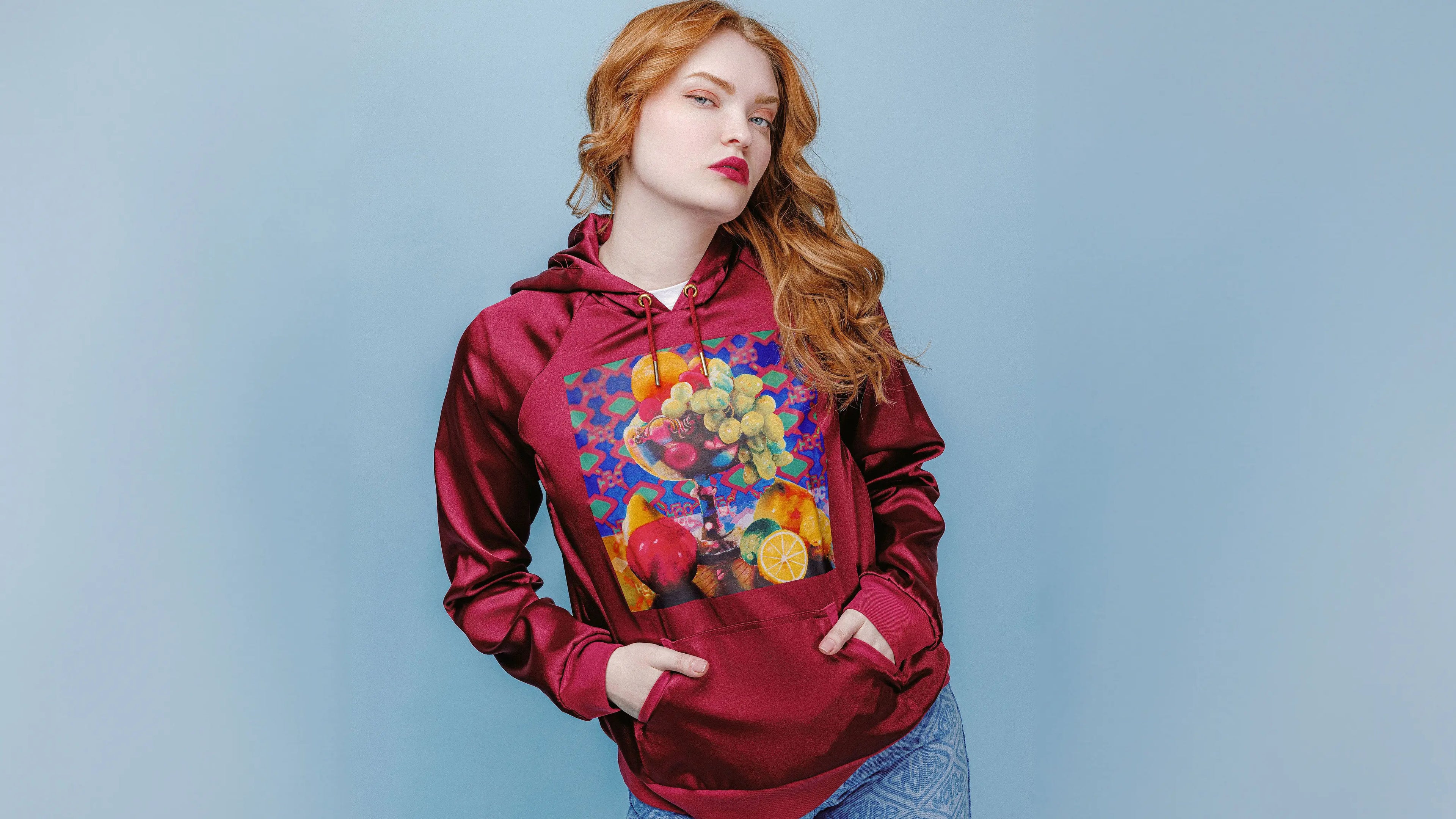 Model wearing the Gabriela Satin Hoodie from House of Caswell's Still Life collection, featuring a rich merlot satin finish and the original Still Life pastel artwork printed on the front against a soft blue studio backdrop.