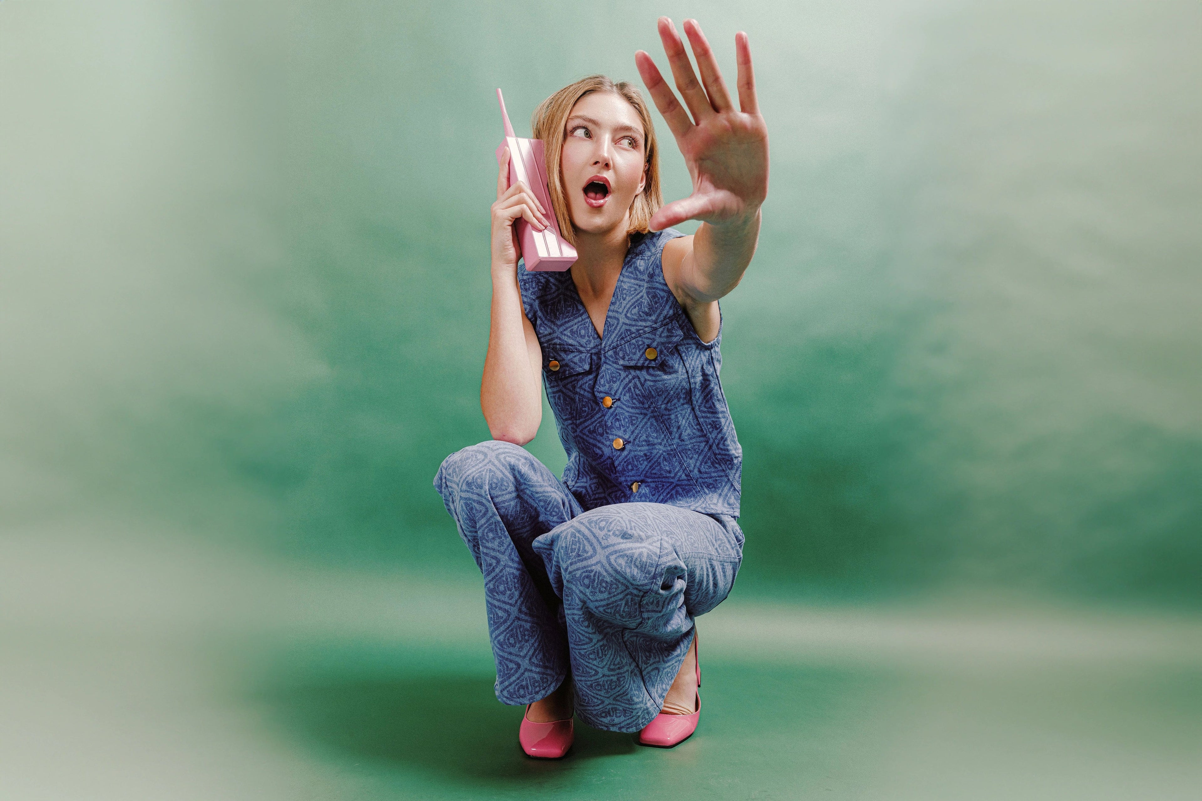 Model wearing the Adriana printed denim vest and matching wide-leg jeans, crouched against a soft green backdrop, holding a pink retro phone and reaching forward, showcasing the bleached monogram denim pattern and gold button details.