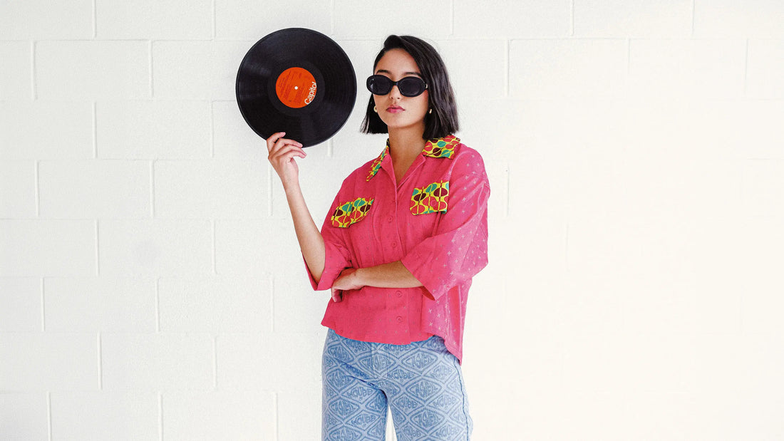 A model wearing the House of Caswell Veronica top and printed denim jeans stands against a white studio wall holding a vinyl record. She wears black sunglasses and glossy pink shoes, blending retro music culture with artful contemporary styling.