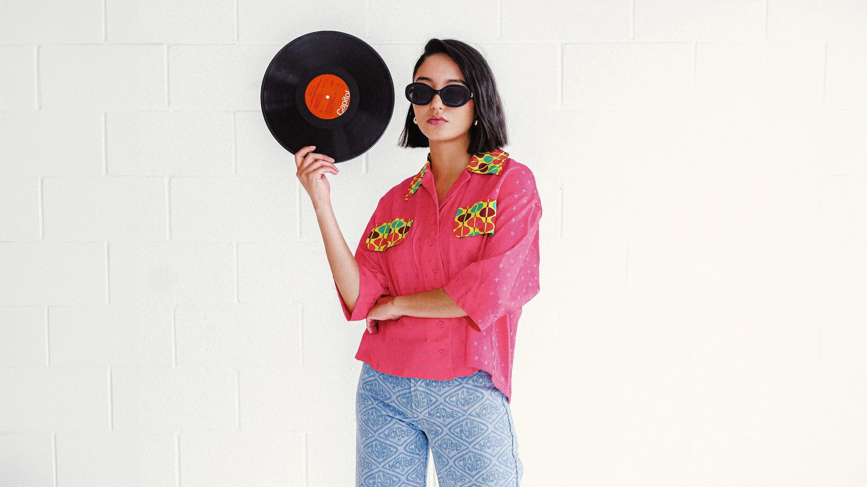 A model wearing the House of Caswell Veronica top and printed denim jeans stands against a white studio wall holding a vinyl record. She wears black sunglasses and glossy pink shoes, blending retro music culture with artful contemporary styling.