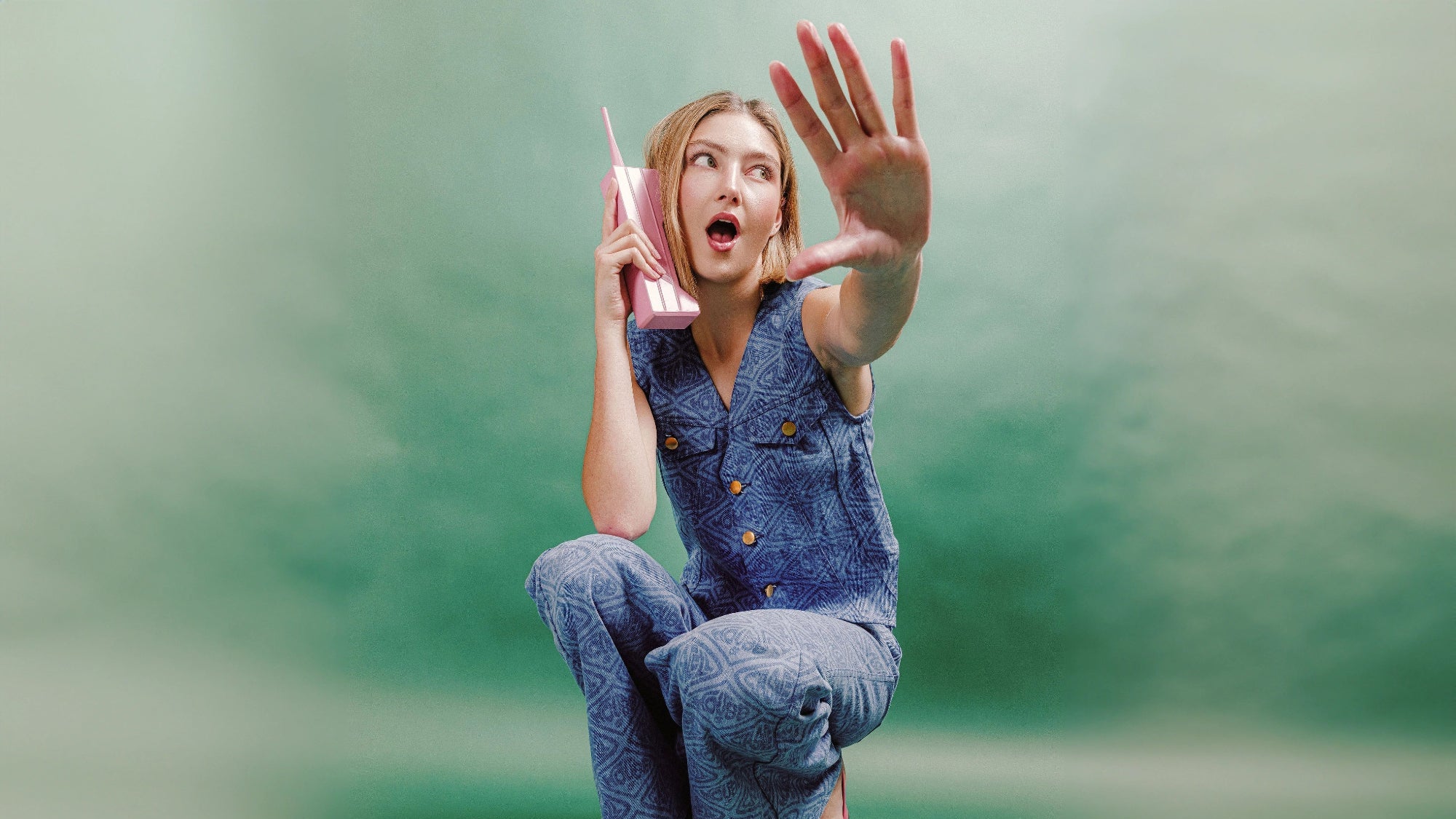 Woman in denim outfit holding a pink phone against a green background