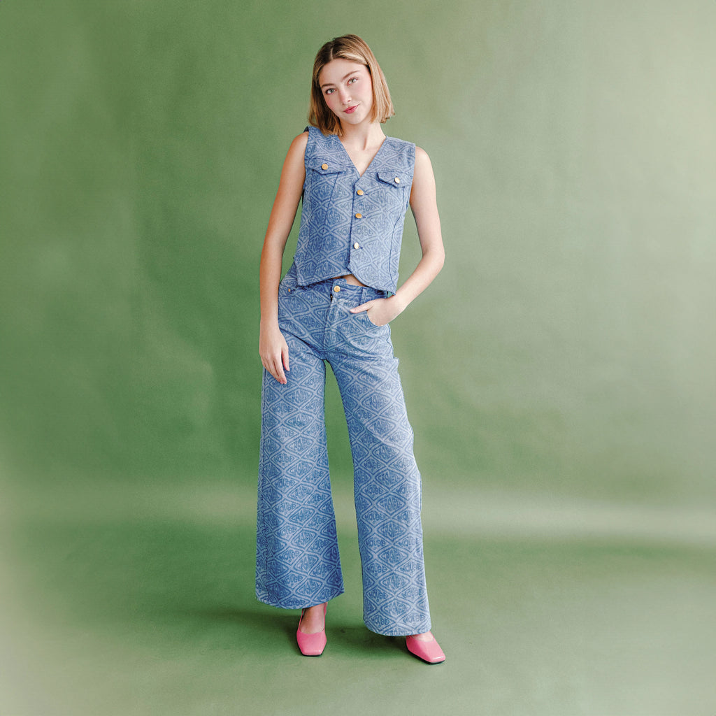 Model wearing a blue patterned sleeveless vest and matching wide-leg pants, standing against a soft green studio backdrop with hands in pockets.