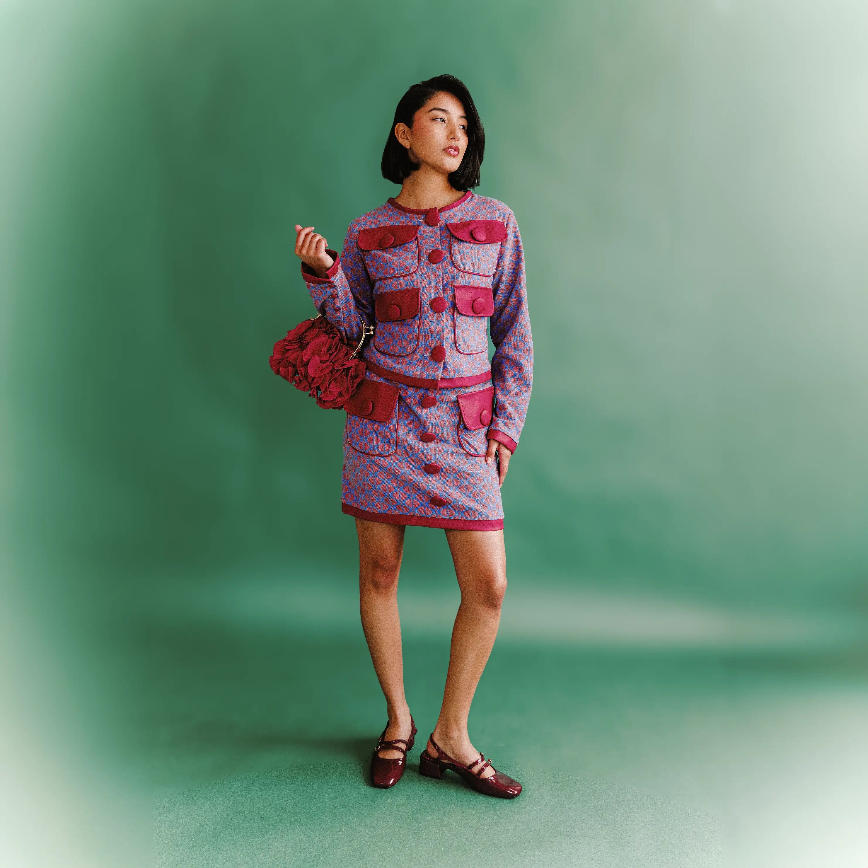 Model wearing the Carly Ensemble by House of Caswell, a patterned cardigan jacket and mini skirt set with red trim and structured pockets, photographed against a green backdrop.