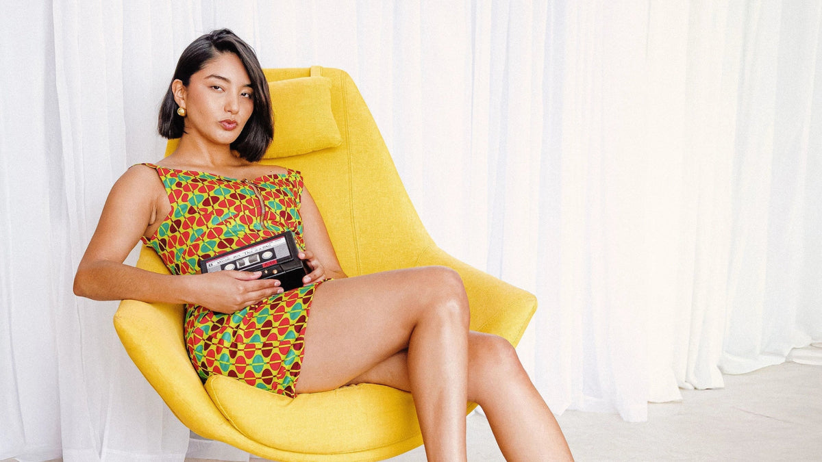 Woman sitting on a yellow chair holding a cassette tape in a room with white curtains.
