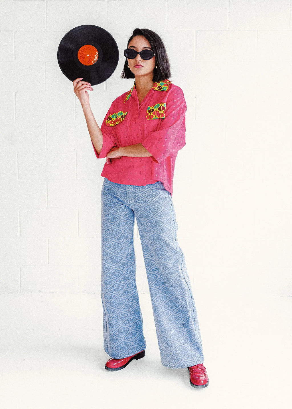 Woman wearing a bright pink printed shirt with and blue patterned pants, holding a vinyl record.