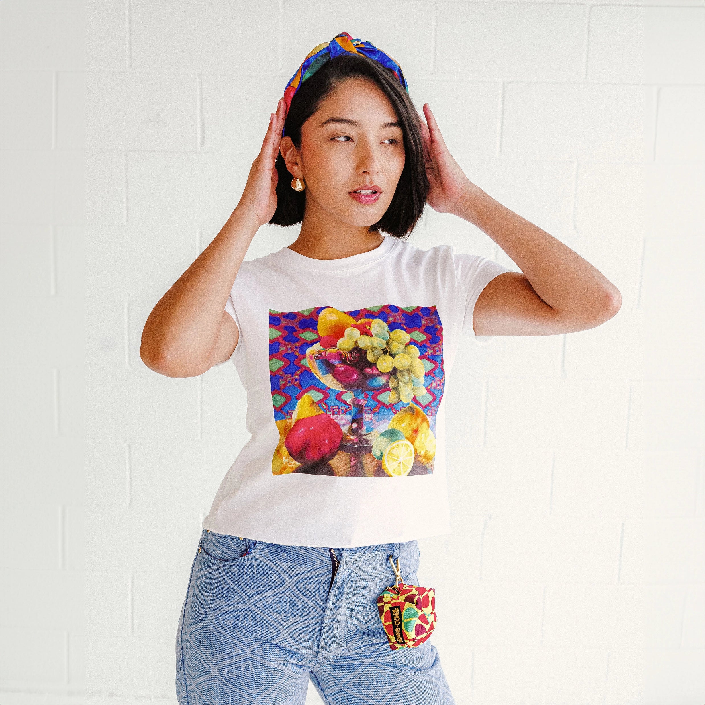 Model wearing a white graphic tee featuring the original Still Life artwork, Adriana printed denim jeans, a Kaitlin charm bag clipped at the waist, and a colorful turban headband.